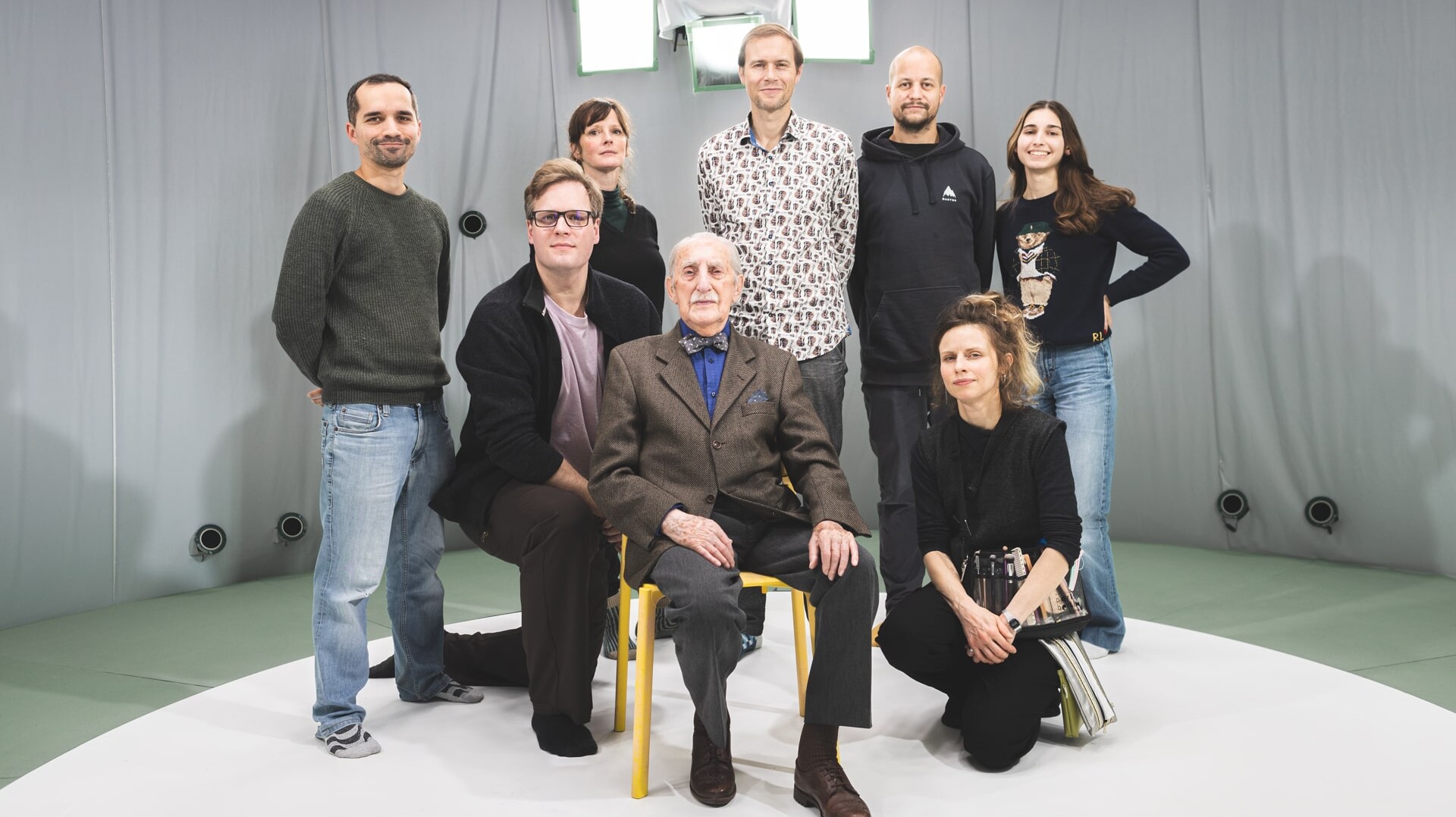 Holocaust survivor Leon Weintraub sits in a yellow chair surrounded by a group of collaborators and filmmakers in the Volucap Studio, with volumetric capture equipment visible in the background.